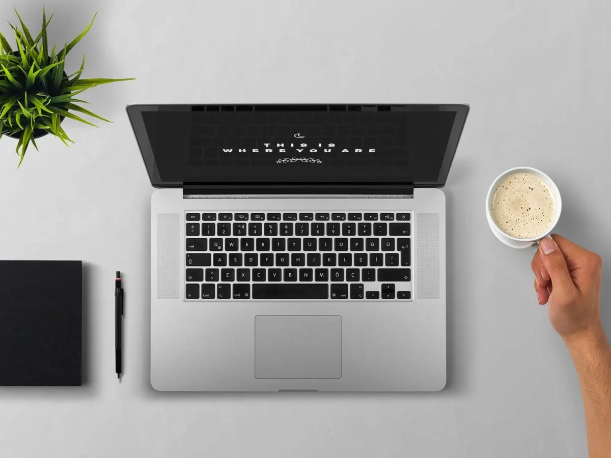 Une photo d'un ordinateur, un cahier, un crayon, un plante et une main tenant une tasse de café
