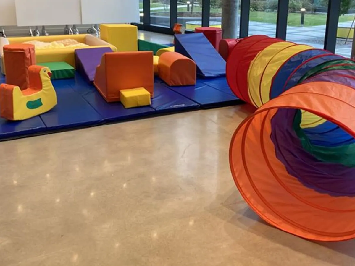 Un espace de jeu pour les enfants d'âge préscolaire, avec divers modules de jeux colorés, dont un tunnel, un tapis coussiné et des blocs colorés.