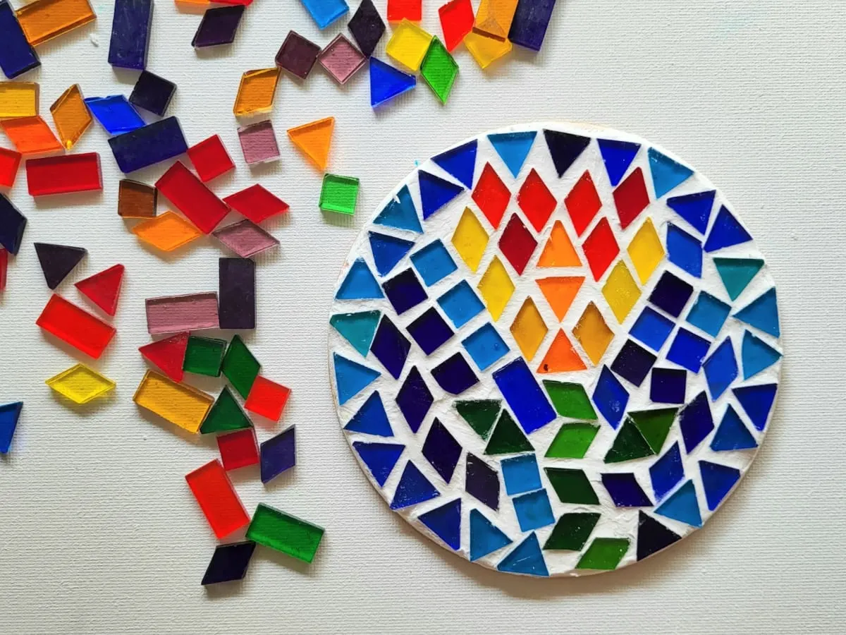 Une mosaïque de forme ronde présente une tulipe dans les tons de jaune, orange et rouge, avec une tige vert pâle et deux feuilles, en vert plus foncé. Elle se détache du fond bleu. À gauche de la mosaïque, plusieurs morceaux de verre, de formes et de couleurs différentes, sont éparpillés.