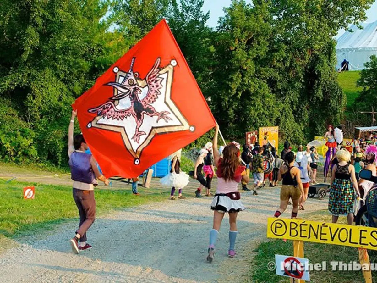 Une foule participe à un événement en plein air sur un chemin de gravier, avec une grande bannière rouge portée à l’avant, entourée de verdure et de chapiteaux en arrière-plan. Un panneau jaune indique la direction des bénévoles.