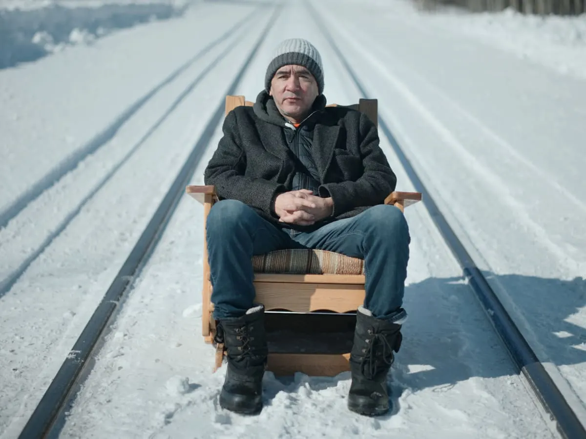 Une personne est assise sur un fauteuil en bois placé au milieu de rails de train recouverts de neige.