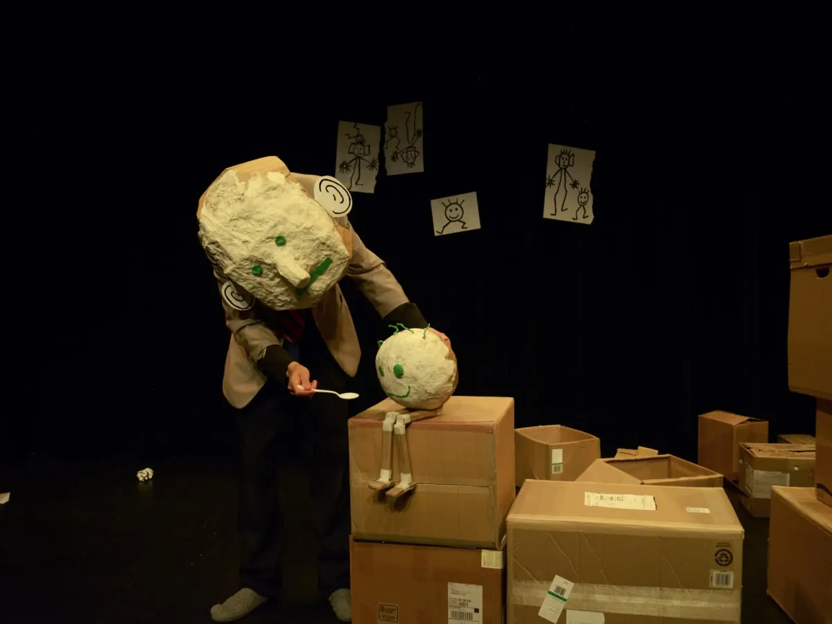 Performance théâtrale avec personnage à tête de papier manipulant un objet sur scène entourée de boîtes en carton.
