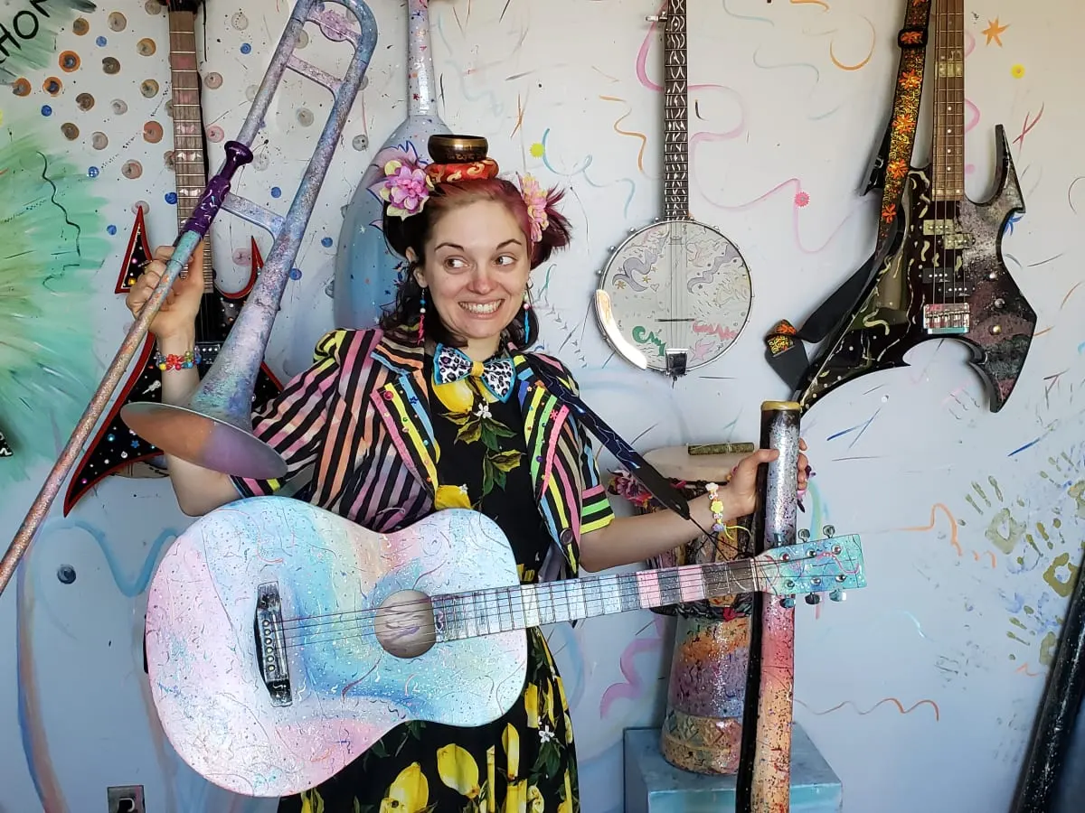 Photo de l'animatrice Laura Ouistiti de la joie qui tient une guitare acoustique colorée devant un mur d'instruments de musique variés.