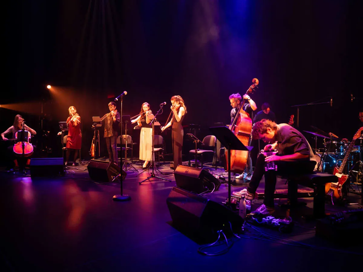 Des musiciennes et musiciens jouent de leur instrument sur scène devant un fond noir.