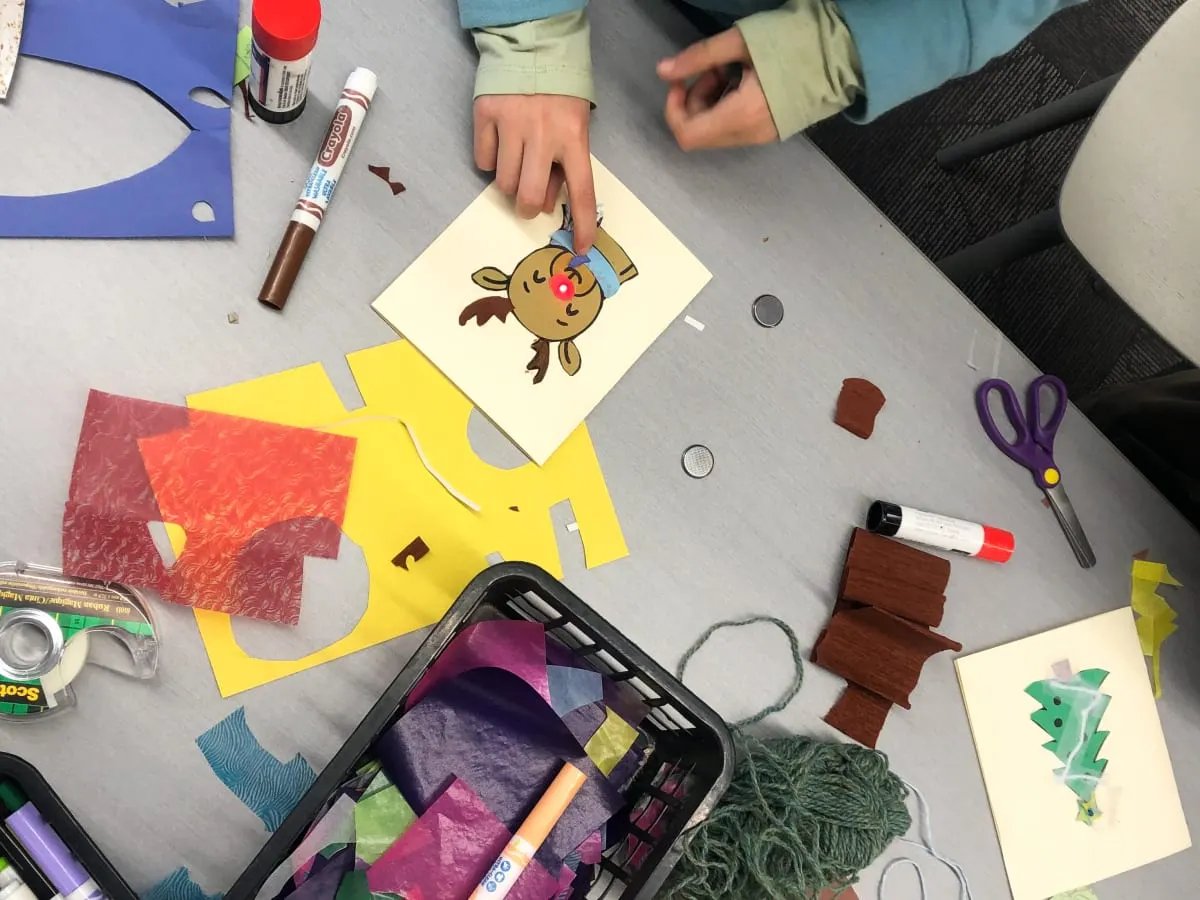 On voit des mains d'enfant qui manipulent du matériel de bricolage, des cartes, cartons de couleur, crayons et colle.