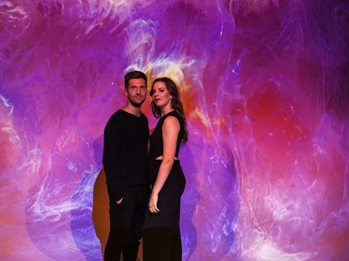 Un homme et une femme vêtus de noir se tiennent devant un fond aux nuances de rose et de lavande.