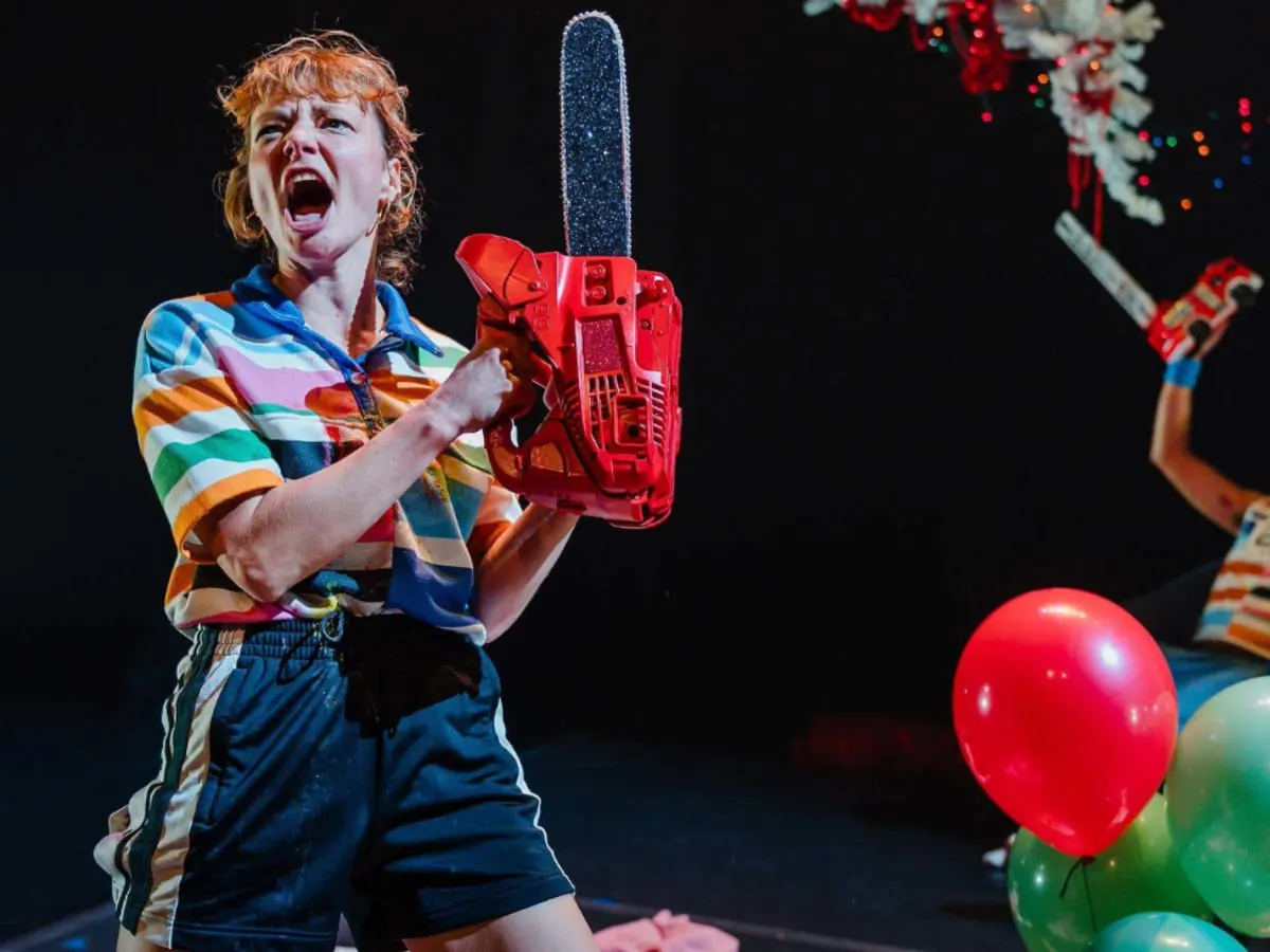 Actrice tenant une tronconneuse en criant. À la droite, des ballons colorés et un acteur qui tient un mini camion de pompier à bout de bras en arrière plan.