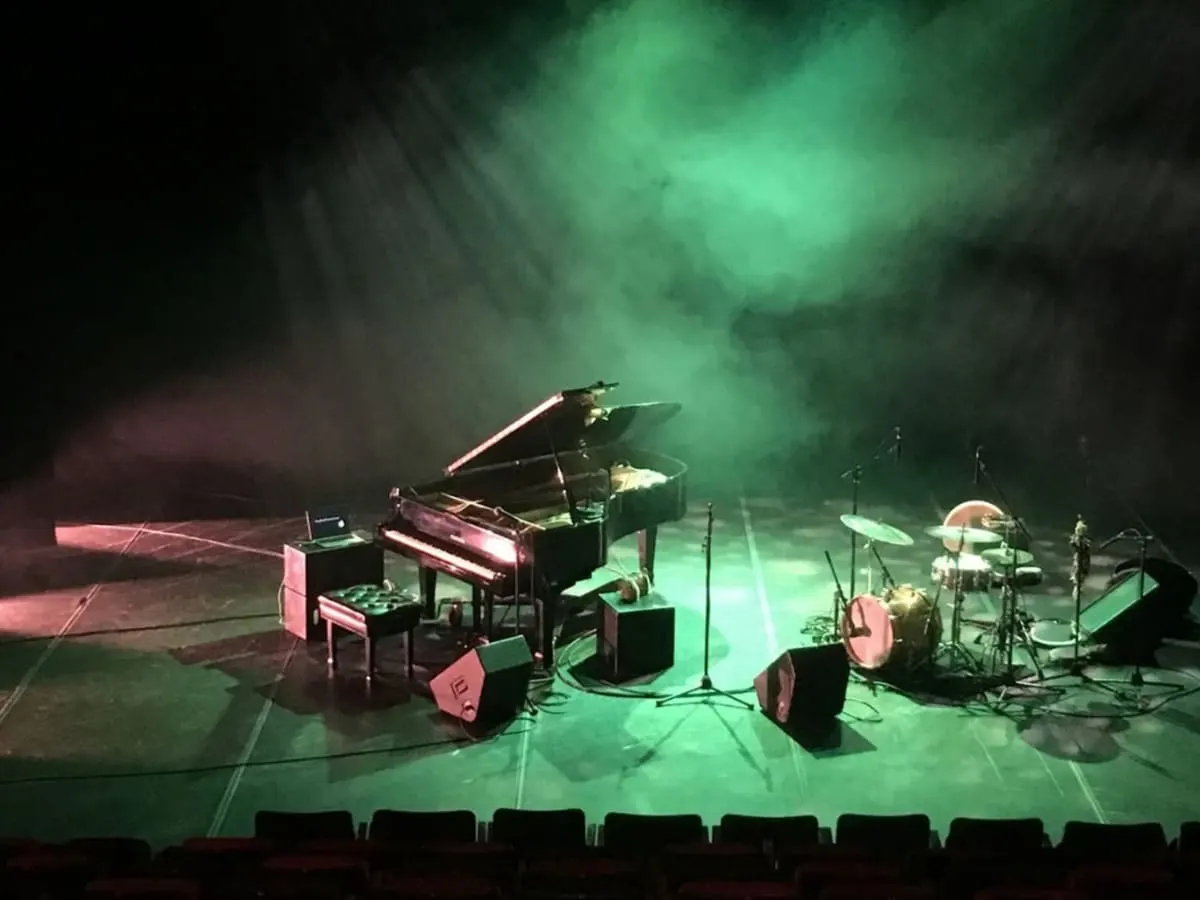 Une scène de spectacle vide avec un piano à queue, une batterie et des amplificateurs sous un éclairage vert et une légère brume.