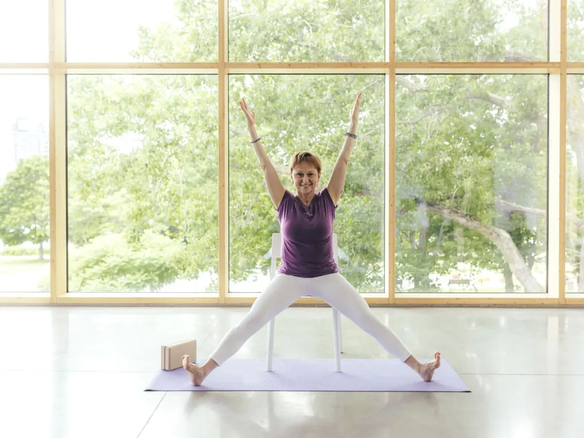 Une femme souriante qui porte un chandail mauve et un pantalon blanc assise sur une chaise avec les bras étendus vers le plafond et les jambes étendues aux côtés.